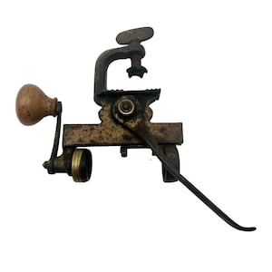 Puede incluir: Una herramienta de metal antigua con un mango de madera y una palanca. La herramienta tiene una abrazadera y un mecanismo de engranaje. Es probable que se use para manualidades o carpintería.