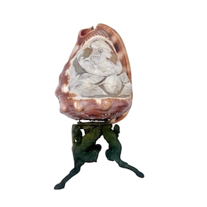 May include: A large, ornate seashell sculpture. The shell is a mix of brown and cream colors, with a carved relief of a classical figure. It rests on a dark green, bronze-toned stand with three supporting figures. The sculpture is set against a white background.