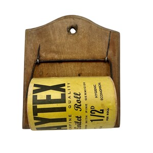 May include: Vintage wooden toilet paper holder with a roll of "NYTEX" toilet paper. The holder has a rectangular backboard with a curved top and a hole for hanging. The toilet paper roll is yellow with black text.