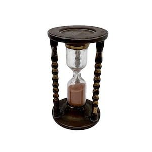 Op de afbeelding: Een houten zandloper met een messing voet en een glazen bol gevuld met bruin zand. De zandloper is ongeveer 25 cm hoog.