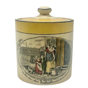 May include: A vintage ceramic jar with a yellow lid and a hand-painted scene of a woman and two children selling milk. The jar is decorated with a black and white border and the text "Cries of London" in a decorative font.