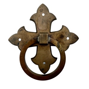 May include: A brass door knocker in the shape of a cross with a ring pull. The cross has a decorative design with four points and the ring is attached to the center of the cross.