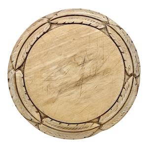 May include: A circular light-coloured wooden board featuring a smooth, scratched central surface surrounded by a carved border of repeating leaf-like patterns. The natural wood grain is visible throughout the piece, which has a flat, round shape.