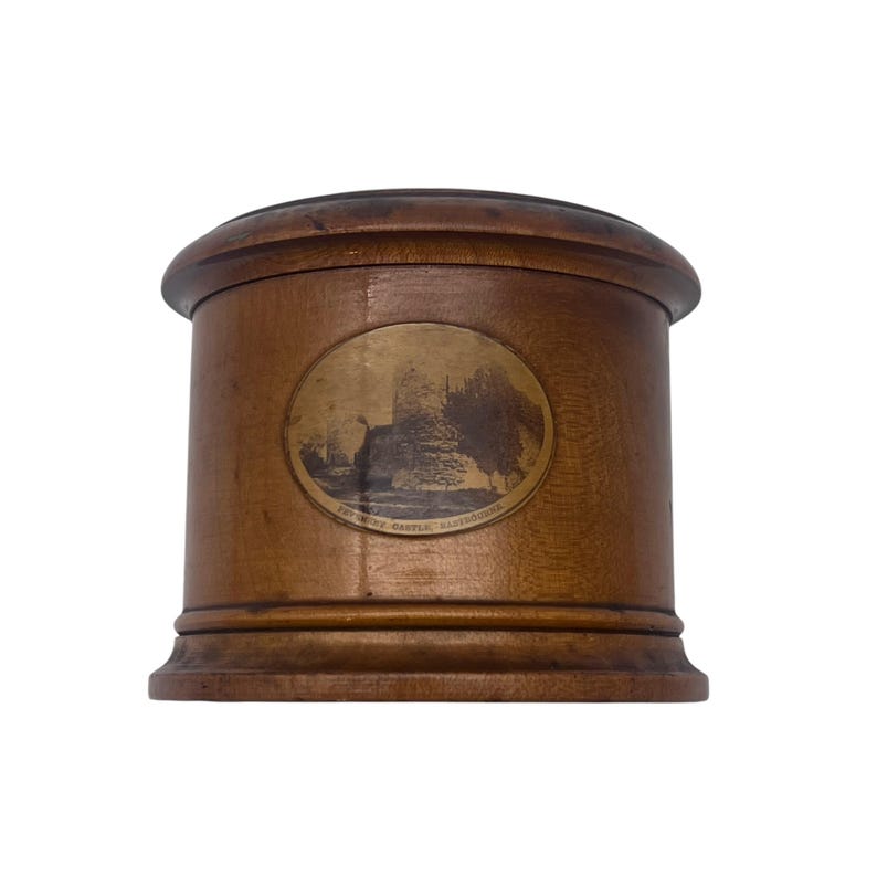 May include: A wooden cylindrical box with a round, brass-colored label featuring a black and white image of a castle and the text "Framlingham Castle, Suffolk".
