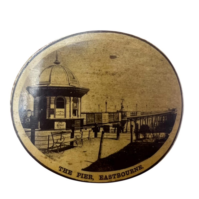 May include: A vintage postcard featuring a black and white photograph of a pier in Eastbourne, England. The pier has a domed building at the beginning of the pier and the text "THE PIER, EASTBOURNE" is printed at the bottom of the postcard.