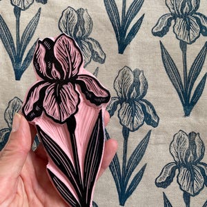 Op de afbeelding: Een roze en zwarte irisbloemstempel met een gedetailleerd gesneden ontwerp, vastgehouden in een hand. De achtergrond toont een stof met een herhalend patroon van blauwe irisbloemen.