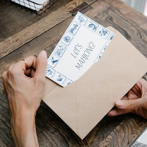 Pu&ograve; includere: Una busta di carta kraft contiene una carta con il testo "Let's MAHJONG!". La carta ha uno sfondo bianco con un bordo blu di illustrazioni decorative. La scena &egrave; ambientata su un tavolo di legno.