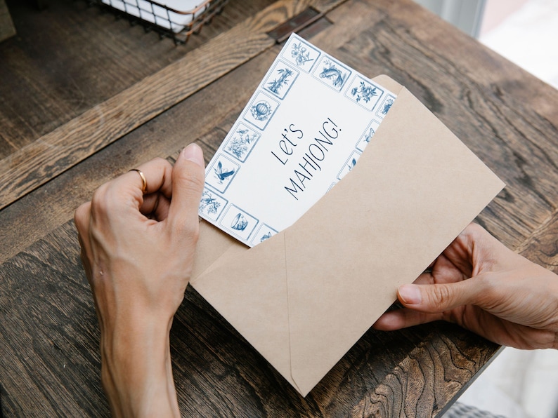 Pu&ograve; includere: Una busta di carta kraft contiene una carta con il testo "Let's MAHJONG!". La carta ha uno sfondo bianco con un bordo blu di illustrazioni decorative. La scena &egrave; ambientata su un tavolo di legno.