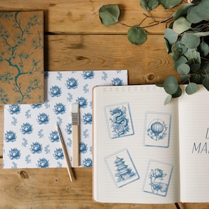 Pu&ograve; includere: Un taccuino aperto con il testo "Let's MAHJONG!" accanto a quattro carte illustrate in blu e bianco. Sono visibili anche un taccuino a spirale e carta fantasia con disegni floreali blu, insieme a un pennello e una penna.