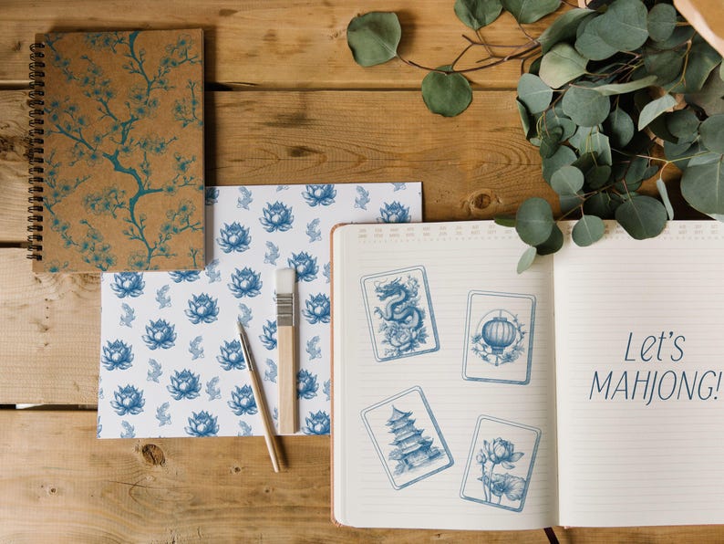 Pu&ograve; includere: Un taccuino aperto con il testo "Let's MAHJONG!" accanto a quattro carte illustrate in blu e bianco. Sono visibili anche un taccuino a spirale e carta fantasia con disegni floreali blu, insieme a un pennello e una penna.