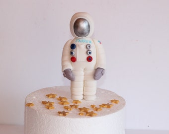 May include: A white fondant astronaut cake topper with the name "Jasper" on the chest. The astronaut is standing on a white cake with gold star sprinkles.