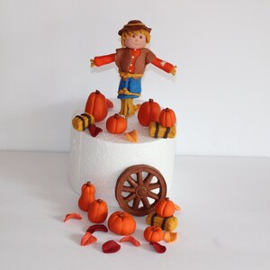 May include: A scarecrow cake topper with a brown hat, brown shirt, blue pants, and straw arms and legs. The topper is surrounded by orange pumpkins, brown hay bales, and brown leaves. The cake topper is on a white cake stand with a brown wooden wheel.