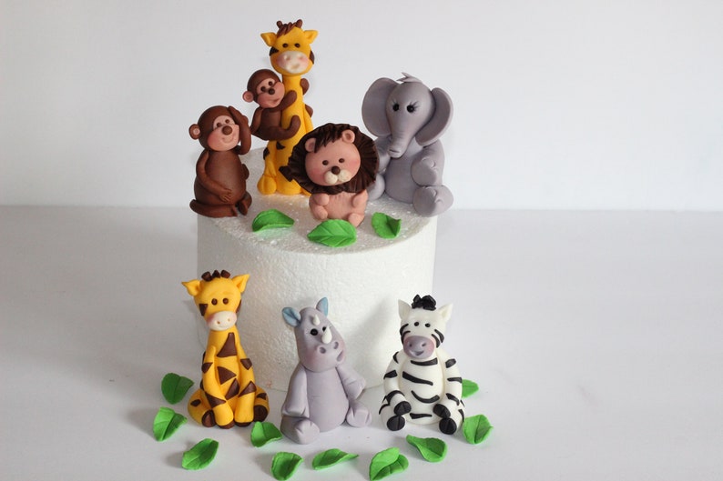 May include: A set of 8 fondant animal cake toppers, including a giraffe, monkey, lion, elephant, rhino, zebra, and two baby giraffes. The toppers are on a white cake with green fondant leaves.