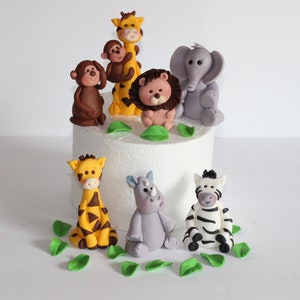 May include: A set of 8 fondant animal cake toppers, including a giraffe, lion, monkey, elephant, zebra, rhino, and two baby giraffes. The toppers are decorated with details like stripes, spots, and facial features. The set also includes green fondant leaves.