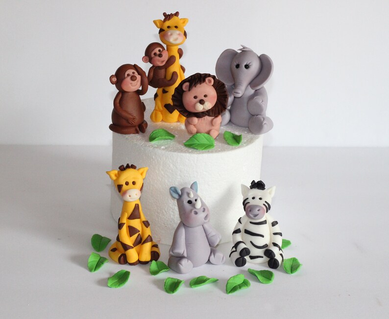 May include: A set of 8 fondant animal cake toppers, including a giraffe, lion, monkey, elephant, zebra, rhino, and two baby giraffes. The toppers are decorated with details like stripes, spots, and facial features. The set also includes green fondant leaves.