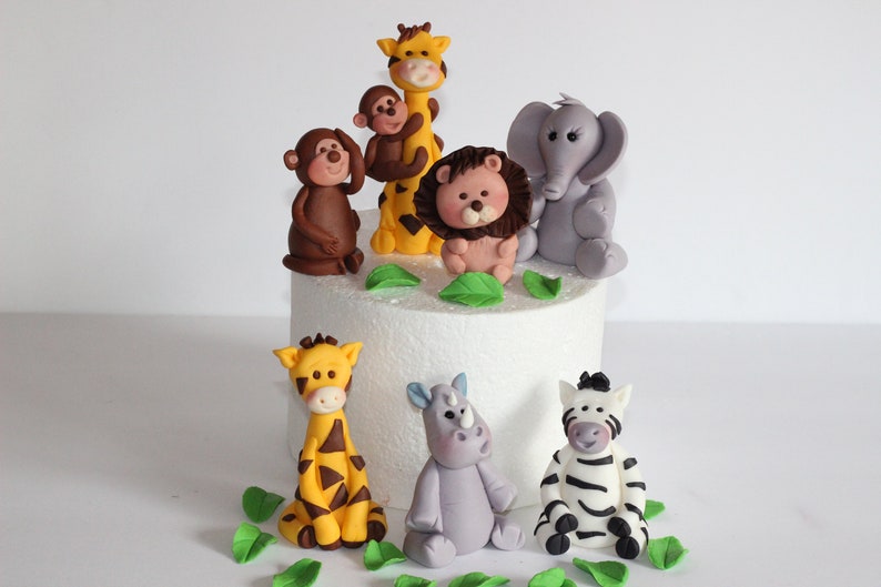 May include: A set of seven fondant animal cake toppers, including a giraffe, lion, elephant, monkey, rhino, zebra, and another giraffe. The toppers are all in various shades of brown, yellow, grey, and white. The toppers are sitting on a white cake.