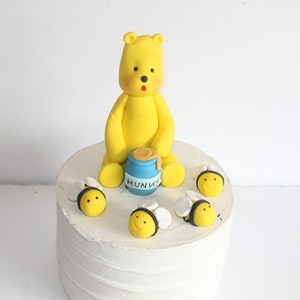 May include: A yellow fondant bear holding a blue honey pot with the word "Hunny" on it. The bear is surrounded by five yellow fondant bees with black stripes on a white frosted cake.
