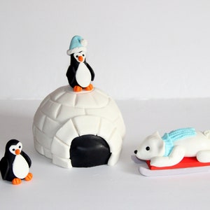 May include: A white and black penguin figurine wearing a blue hat sits on top of a white igloo with a black entrance. A white polar bear figurine wearing a blue scarf is lying on a red sled.