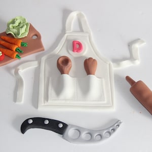 May include: A white fondant apron with a pink letter D, two pockets, and a wooden spoon.  There is a rolling pin, a knife, and a cutting board with fondant carrots, tomatoes, and lettuce.