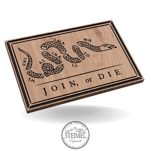 Könnte beinhalten: Rechteckiges Holzschild mit schwarzem Rand und der Aufschrift "JOIN, or DIE". Es zeigt ein segmentiertes Schlangenmuster mit Initialen. Das Holz hat eine natürliche Maserung, und der Text ist eingraviert.