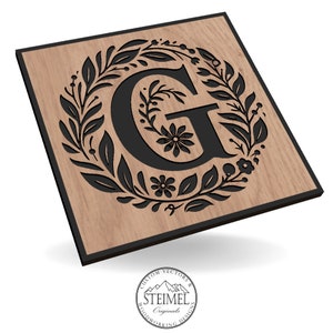 May include: A wooden sign with a black border and a floral wreath design surrounding the letter "G". The wreath is made of leaves and flowers. The sign is made of wood and has a natural wood grain.