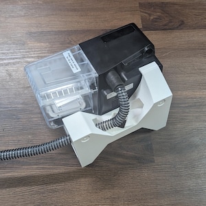 May include: A black and clear plastic device with a white support structure. A flexible, grey and black striped tube extends from the device. The device is on a wooden surface.
