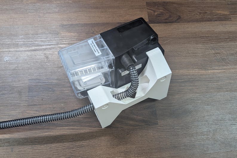 May include: A black and clear plastic device with a white support structure. A flexible, grey and black striped tube extends from the device. The device is on a wooden surface.