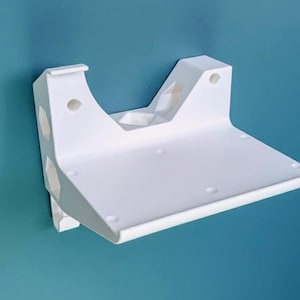 May include: A white wall-mounted shelf with a curved front edge and a flat surface. The shelf has a bracket with several holes for mounting. The shelf is made of a solid material and is against a teal wall.