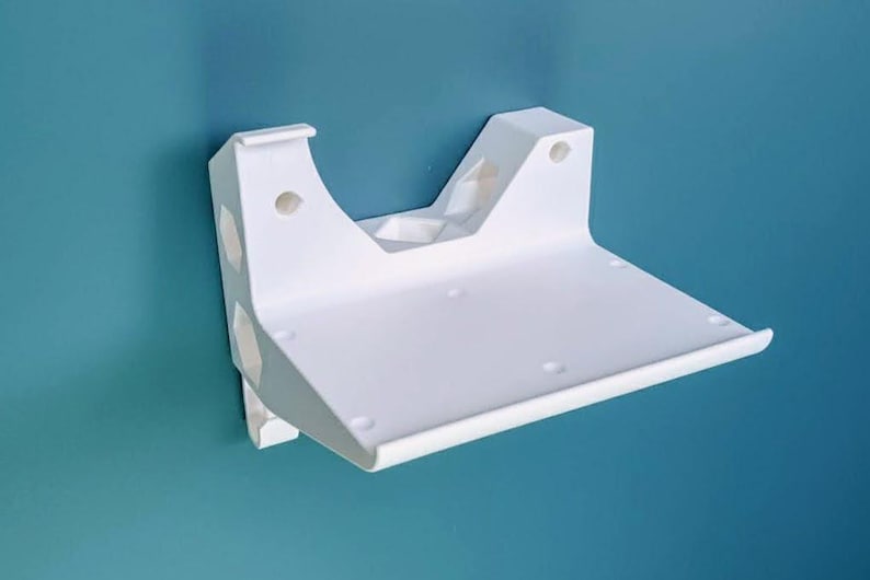 May include: A white wall-mounted shelf with a curved front edge and a flat surface. The shelf has a bracket with several holes for mounting. The shelf is made of a solid material and is against a teal wall.