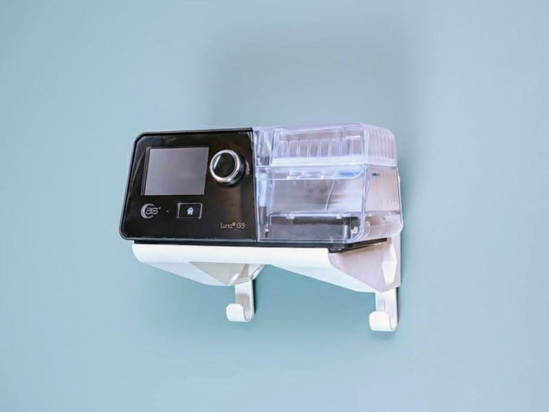 May include: A black and clear medical device with a digital display and a water reservoir. The device is mounted on a white wall bracket. The device has the text "Luna G3".