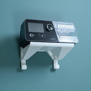 May include: A black and clear medical device with a digital display and control knob, mounted on a white bracket. The device is labelled "Luno G3" and is against a teal wall.