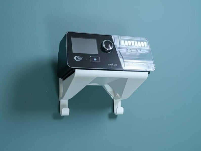 May include: A black and clear medical device with a digital display and control knob, mounted on a white bracket. The device is labelled "Luno G3" and is against a teal wall.