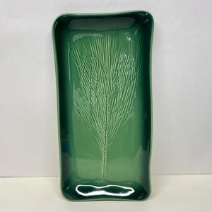 May include: A rectangular, emerald green ceramic tray with rounded corners. The tray features a raised, textured design of a stylized fern frond in the center. The glossy finish enhances the depth of the green color. This decorative tray is suitable for serving or display.