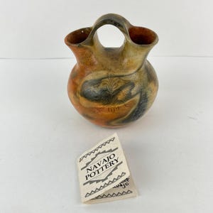 May include: Handmade Navajo pottery vase with a unique handle. The vase features a round body with a flared rim and a handle. The pottery is decorated with earth-tone colors, including orange, brown, and black. The vase has a detailed etched design. A tag reads "Navajo Pottery".