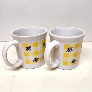 May include: Two white ceramic mugs featuring a yellow and white checkered gingham pattern. Each mug is decorated with small, black line-art illustrations of bees scattered across the checkered surface. Both mugs have a curved handle.