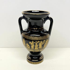 May include: Black ceramic vase with two handles and a gold rim. The vase features a gold Greek key design around the base and a frieze depicting figures in gold. The vase is approximately 8 inches tall.