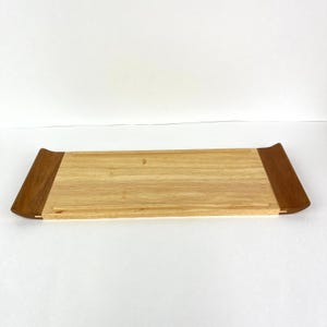 May include: A rectangular wooden serving tray with a light-colored center and darker wood accents on each end. The tray has a slightly curved design and is made of natural wood.