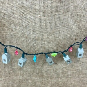 May include: String of decorative lights with small gray house-shaped bulbs and colorful accents. The string includes pink, blue, and green decorative elements. The lights are strung on a dark green wire.
