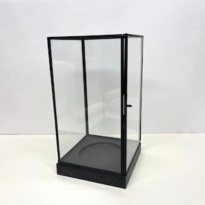 May include: A black rectangular display case with clear glass panels and a hinged door. The case has a black base and frame, and a circular platform inside. The display case is designed to showcase small items.