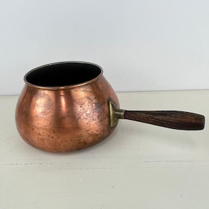 May include: A copper saucepan with a dark interior and a wooden handle. The pot has a rounded body and a small spout. The handle is attached with a brass fitting. The copper has a warm, reddish-brown color.