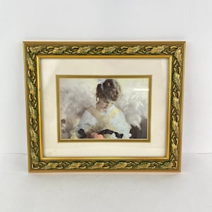 May include: A framed print of a painting depicting a young girl with angel wings holding a small bird in her hands. The frame is gold with a green leaf design.
