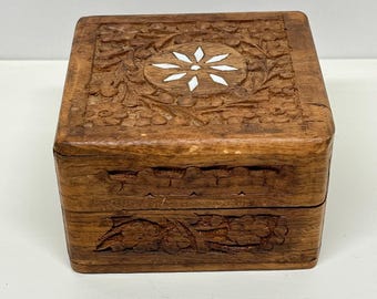 May include: A small, square wooden box with intricate floral carvings. The lid features a central white flower design. The box is made of a warm brown wood and has a hinged construction, suggesting it is for storing small items.