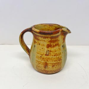 May include: A ceramic pitcher featuring a speckled yellow and brown glaze with a prominent handle and a pouring spout. The exterior surface displays a textured finish with vertical indentations along the sides and a mottled color pattern.
