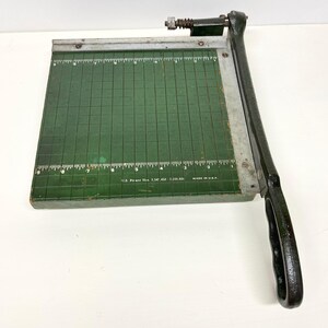 May include: Vintage paper cutter with a dark green cutting surface and a black handle. The cutting surface has grid lines and measurement markings in inches. The blade and handle are black. The text "MADE IN U.S.A." is visible.
