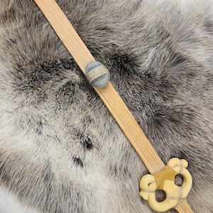 May include: A wooden toy sword with a light brown handle and a decorative, cream-colored hilt. A gray stone is attached to the handle. The sword is lying on a gray and white fur-like surface.