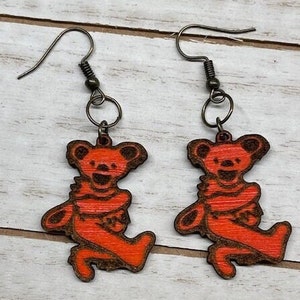 May include: A pair of orange wood earrings featuring a cartoon bear design. The earrings have a simple hook closure.