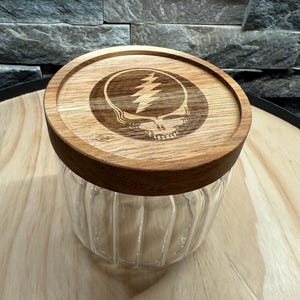 May include: A clear glass jar with a wooden lid. The lid features a laser-etched design of a skull with a lightning bolt. The jar has vertical ridges and sits on a wooden surface. The jar is likely for storage.