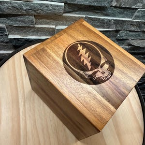 May include: A wooden box with a dark brown finish. The top of the box features a circular design with a skull and lightning bolt graphic. The box is sitting on a light brown surface.