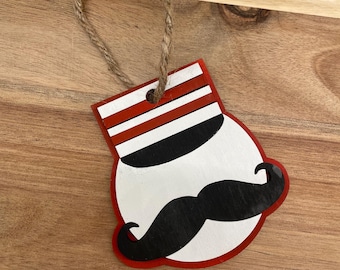 May include: A white wooden ornament shaped like a face with a black mustache and a red and white striped hat. The ornament has a twine loop for hanging.