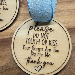 May include: A wooden round sign with the text "please do not touch or kiss your germs are too big for me thank you" with a heart symbol at the bottom. The sign is attached to a blue ribbon with white polka dots.